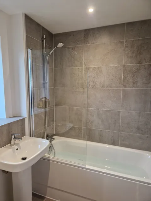 Grey large-format wall tiles over a bath with glass screen, shower and white pedestal basin